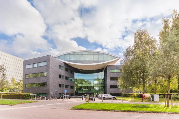 Modern bedrijfsgebouw met een opvallend glazen gevel en een sculptuur van een koe op het gras naast de parkeerplaats.