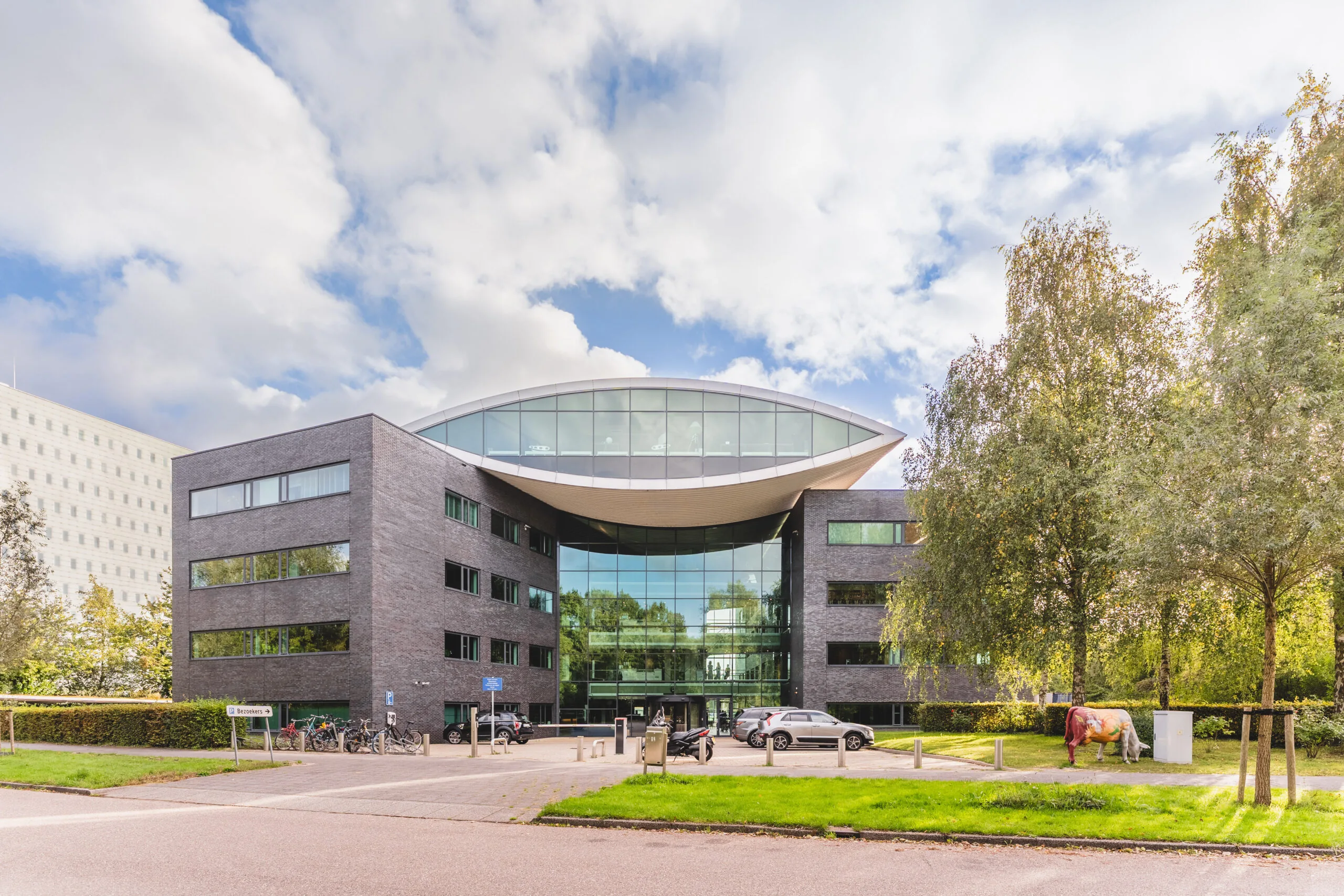 Modern bedrijfsgebouw met een opvallend glazen gevel en een sculptuur van een koe op het gras naast de parkeerplaats.