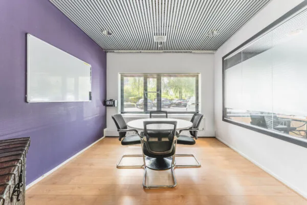 Vergaderruimte met een ronde tafel, vier stoelen, een paarse muur en een groot raam met uitzicht op een parkeerplaats.