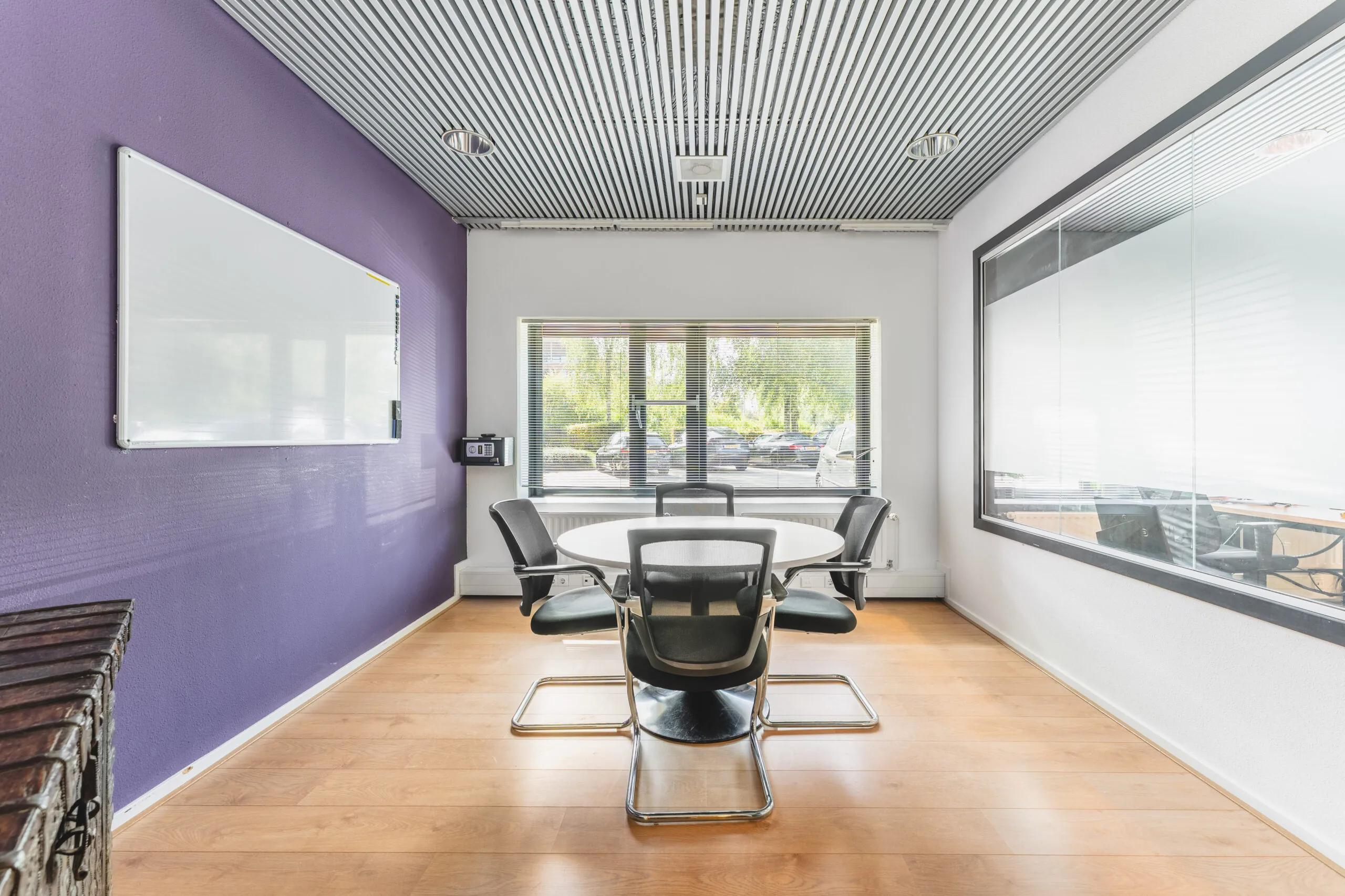 Vergaderruimte met een ronde tafel, vier stoelen, een paarse muur en een groot raam met uitzicht op een parkeerplaats.