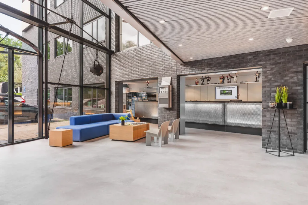 Moderne ontvangstruimte met een blauwe bank, houten tafels en een balie in een gebouw met grote ramen.