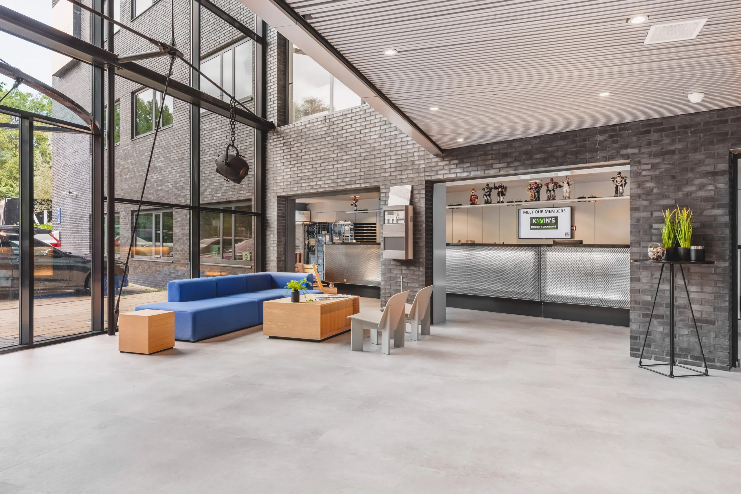 Moderne ontvangstruimte met een blauwe bank, houten tafels en een balie in een gebouw met grote ramen.