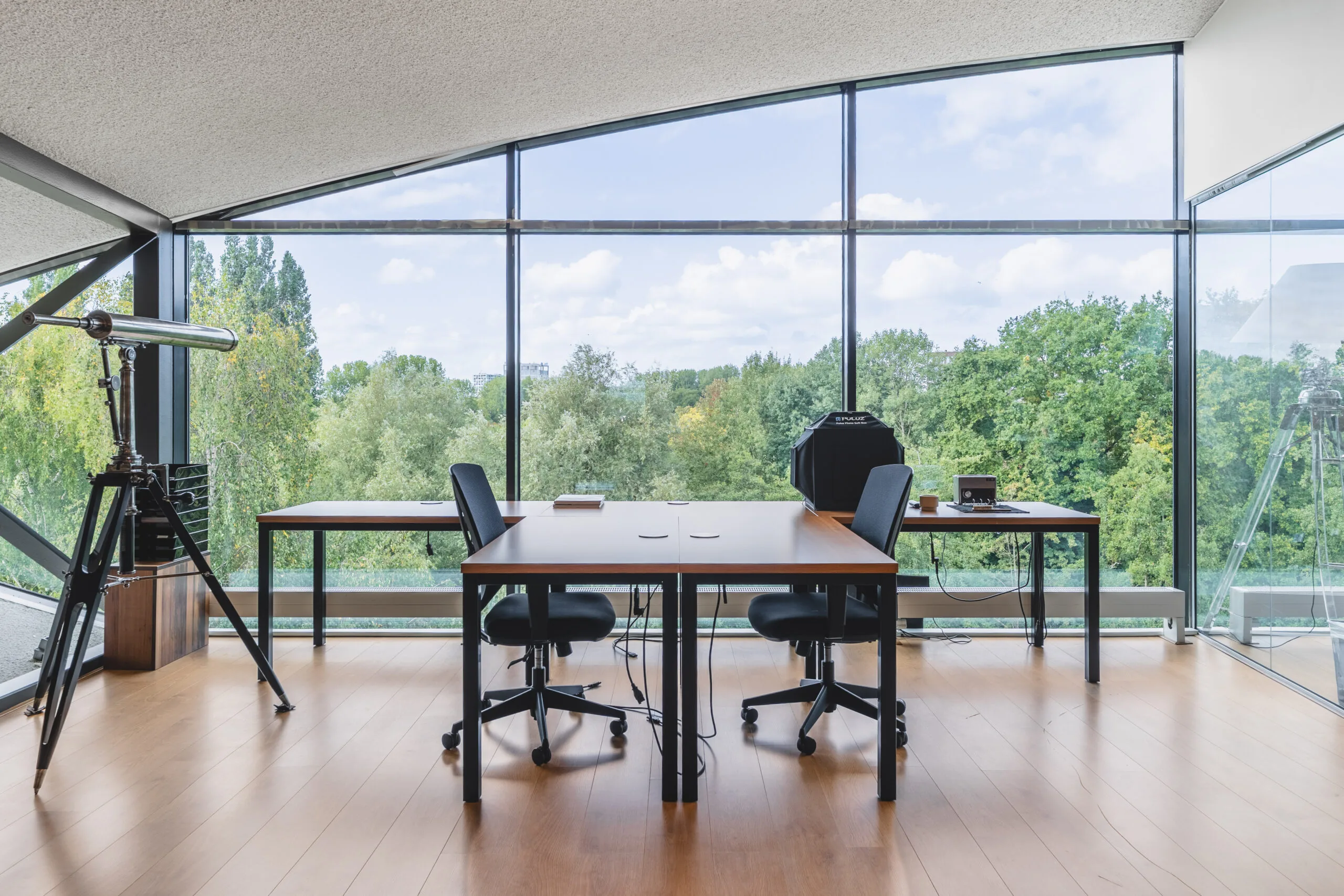 Een moderne kantoorruimte met grote ramen, twee tafels, stoelen en een grote telescoop.