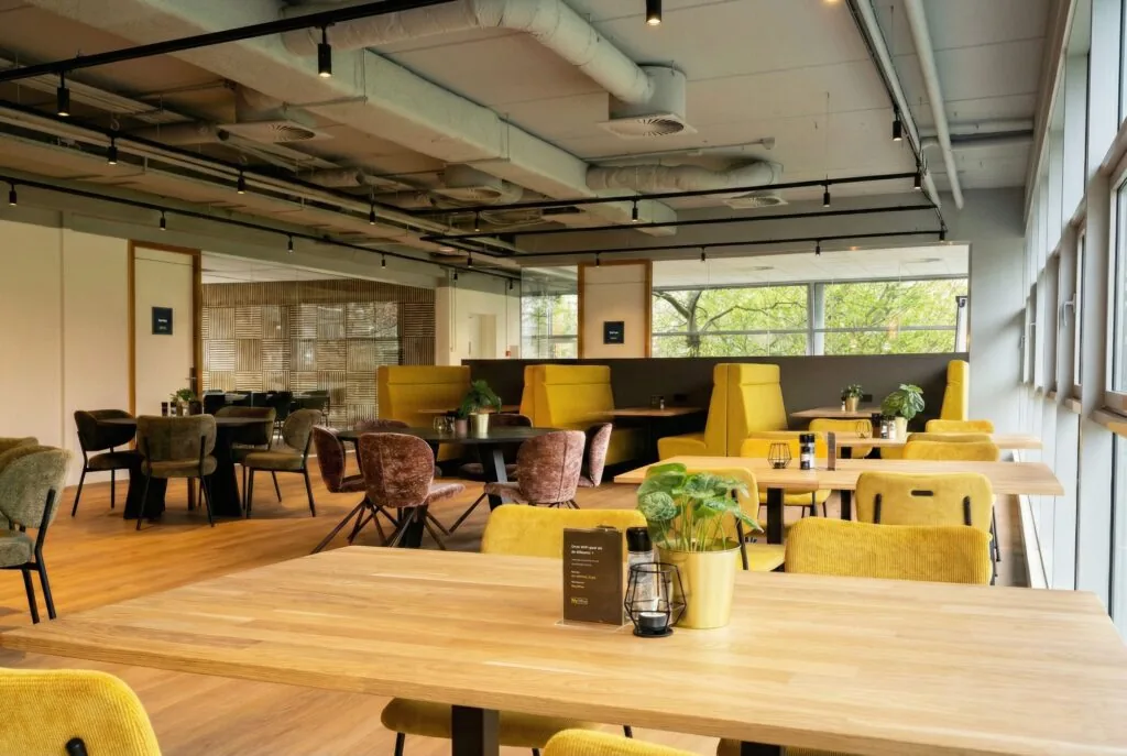 Een modern atrium met houten tafels, kleurrijke stoelen en veel natuurlijk licht.