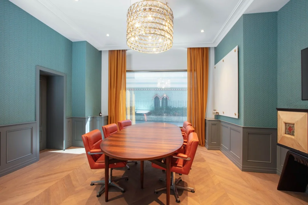 Vergaderruimte in Coolsingel 65 met ovale houten tafel, oranje stoelen, grote kroonluchter en hoge ramen met okergele gordijnen.