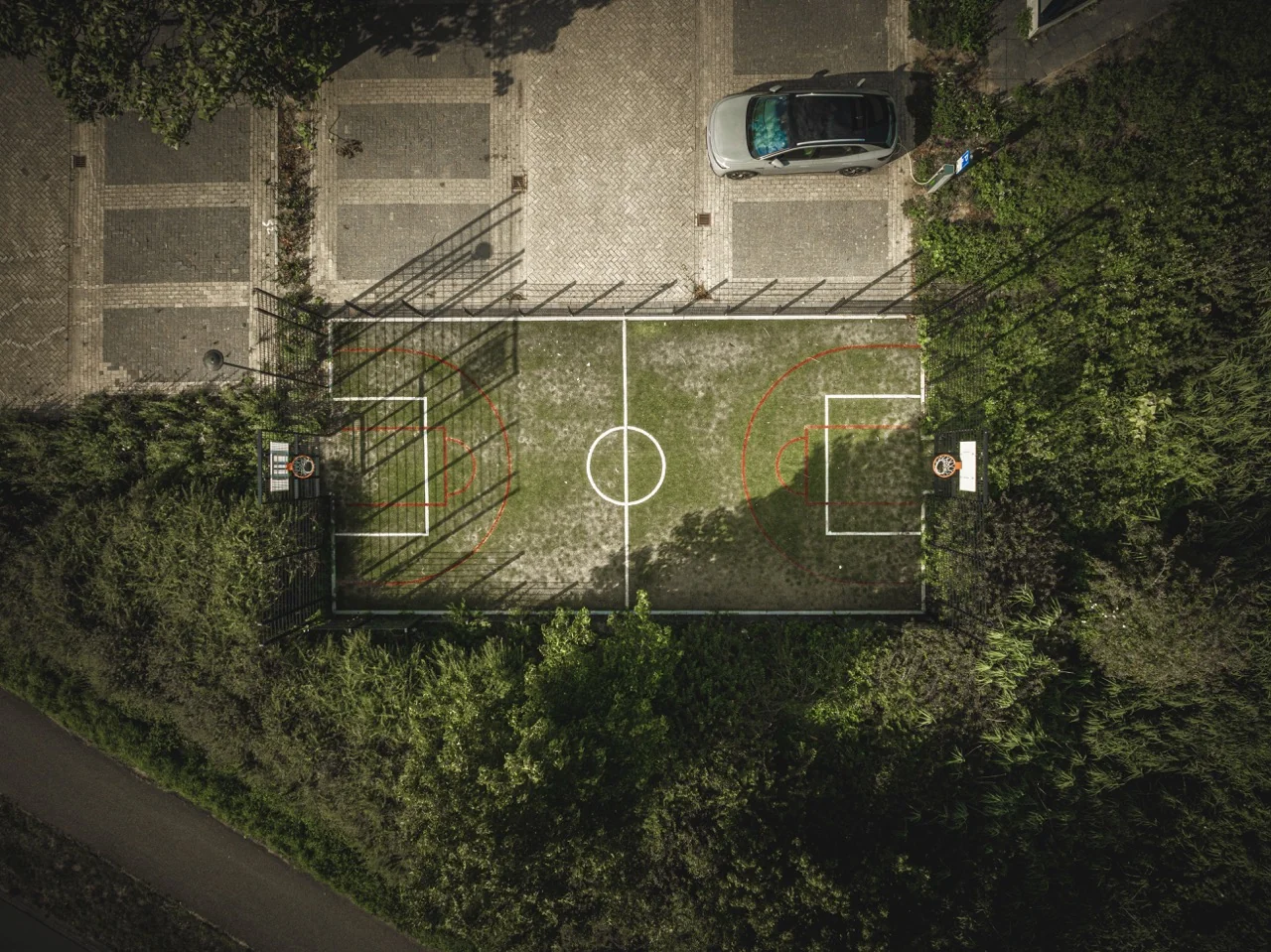 Luchtfoto van een klein sportveld met basketbalringen, omringd door groen en parkeerplaatsen.