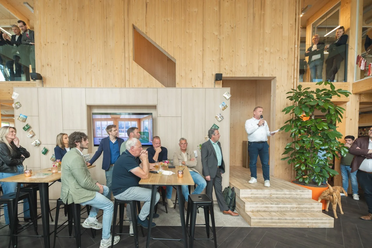 Man spreekt een groep mensen toe met een microfoon in een modern houten interieur tijdens een bijeenkomst.
