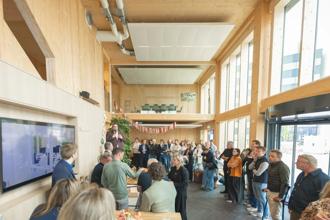 Een groep mensen luistert naar een toespraak tijdens een bijeenkomst in een moderne, houten ruimte met veel natuurlijk licht.