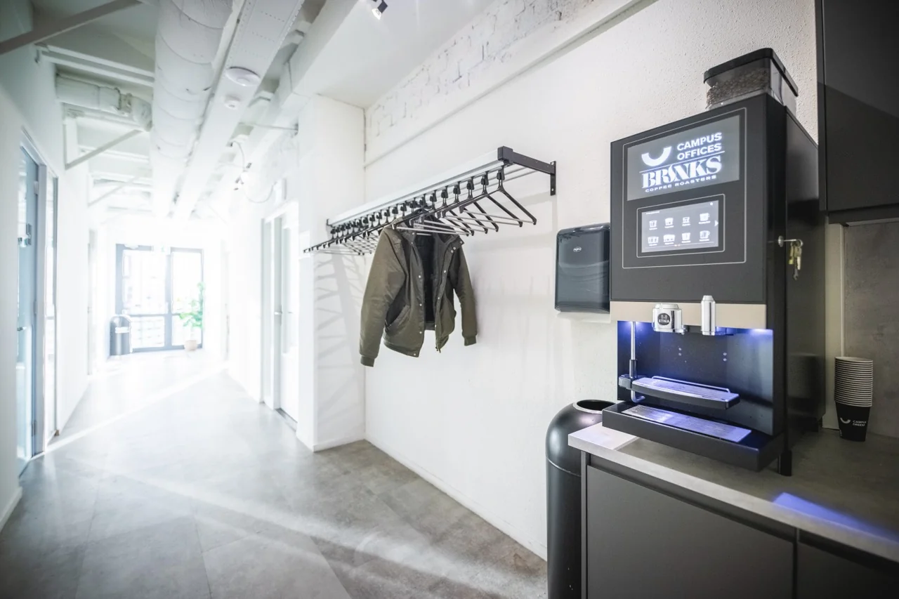 Lichte kantoorhal met een koffieautomaat van Campus Offices BRNKS, een kapstok met jassen en hangers.