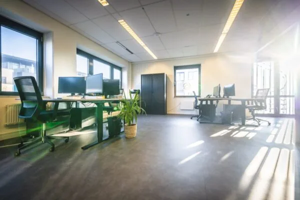 Lichte en moderne kantoorruimte met bureaus, bureaustoelen, computerschermen en grote ramen waar zonlicht naar binnen schijnt.