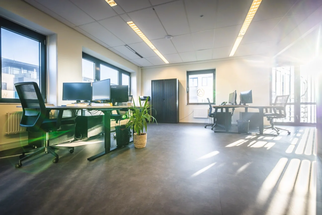 Lichte en moderne kantoorruimte met bureaus, bureaustoelen, computerschermen en grote ramen waar zonlicht naar binnen schijnt.