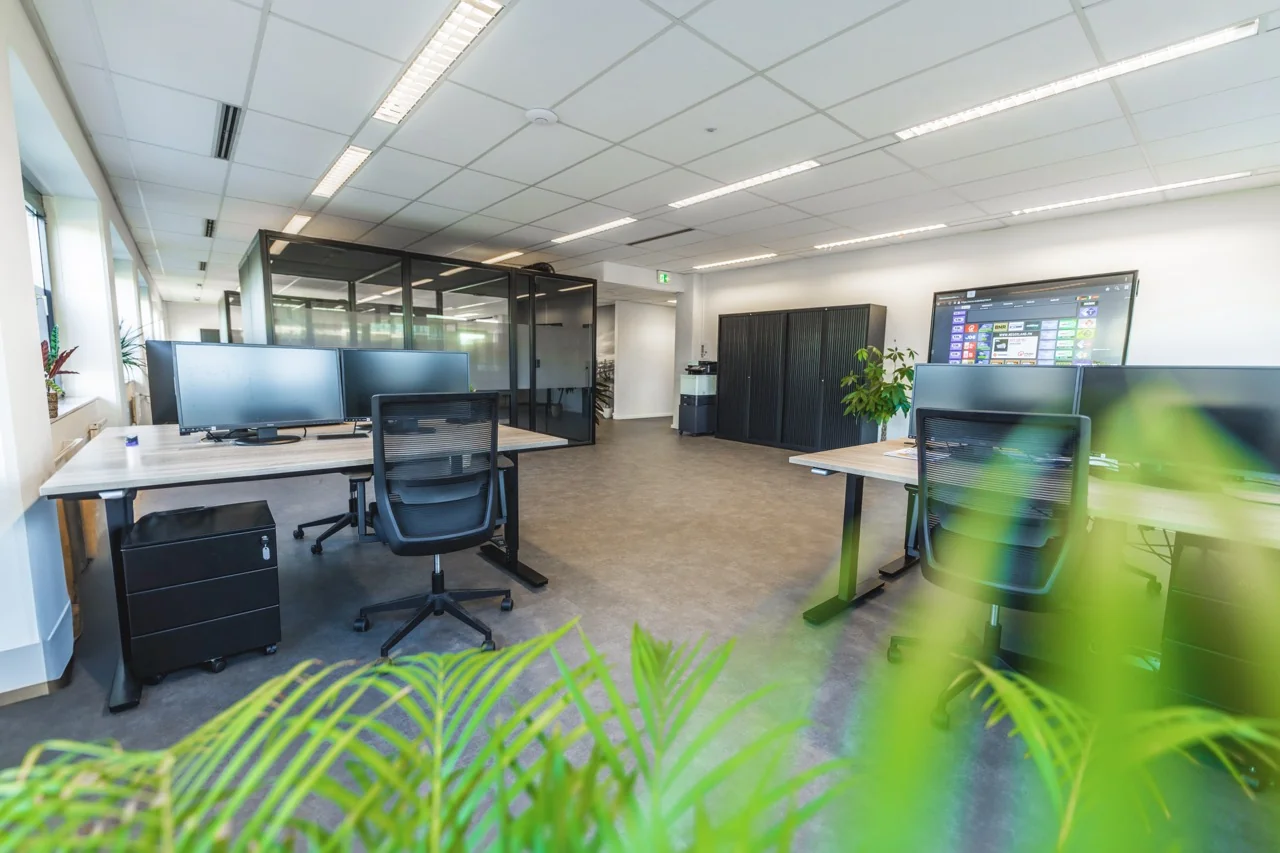 Moderne open kantoorruimte met bureaus, computerschermen, glazen vergaderruimtes en groene planten.