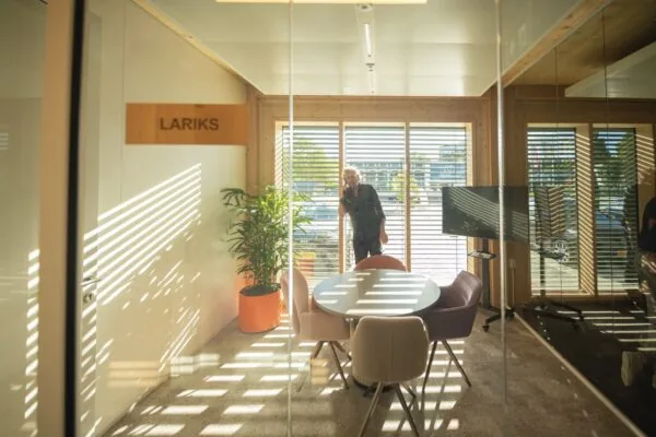 Vergaderruimte genaamd "LARIKS" met ronde tafel, stoelen, grote plant en een man die buiten telefonisch belt achter jaloezieën.