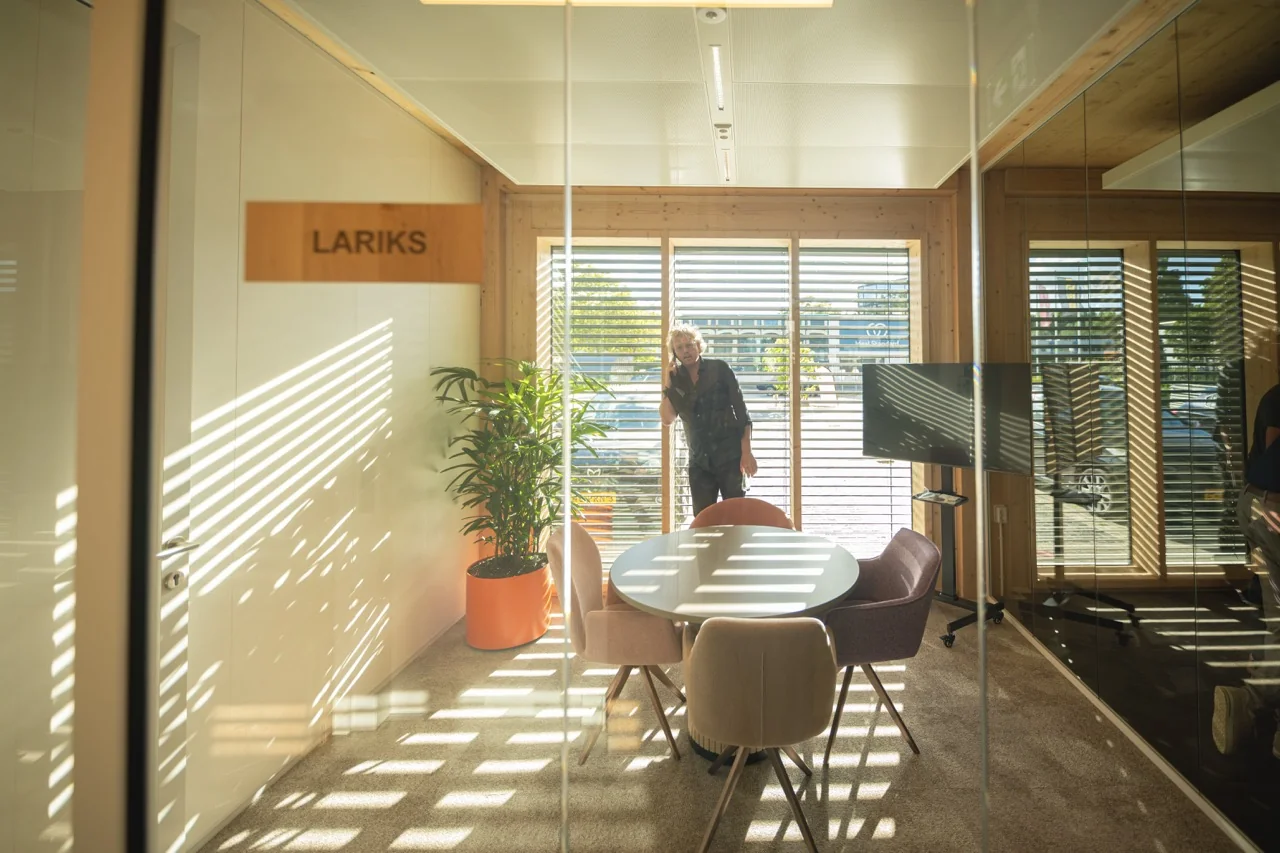 Vergaderruimte genaamd "LARIKS" met ronde tafel, stoelen, grote plant en een man die buiten telefonisch belt achter jaloezieën.