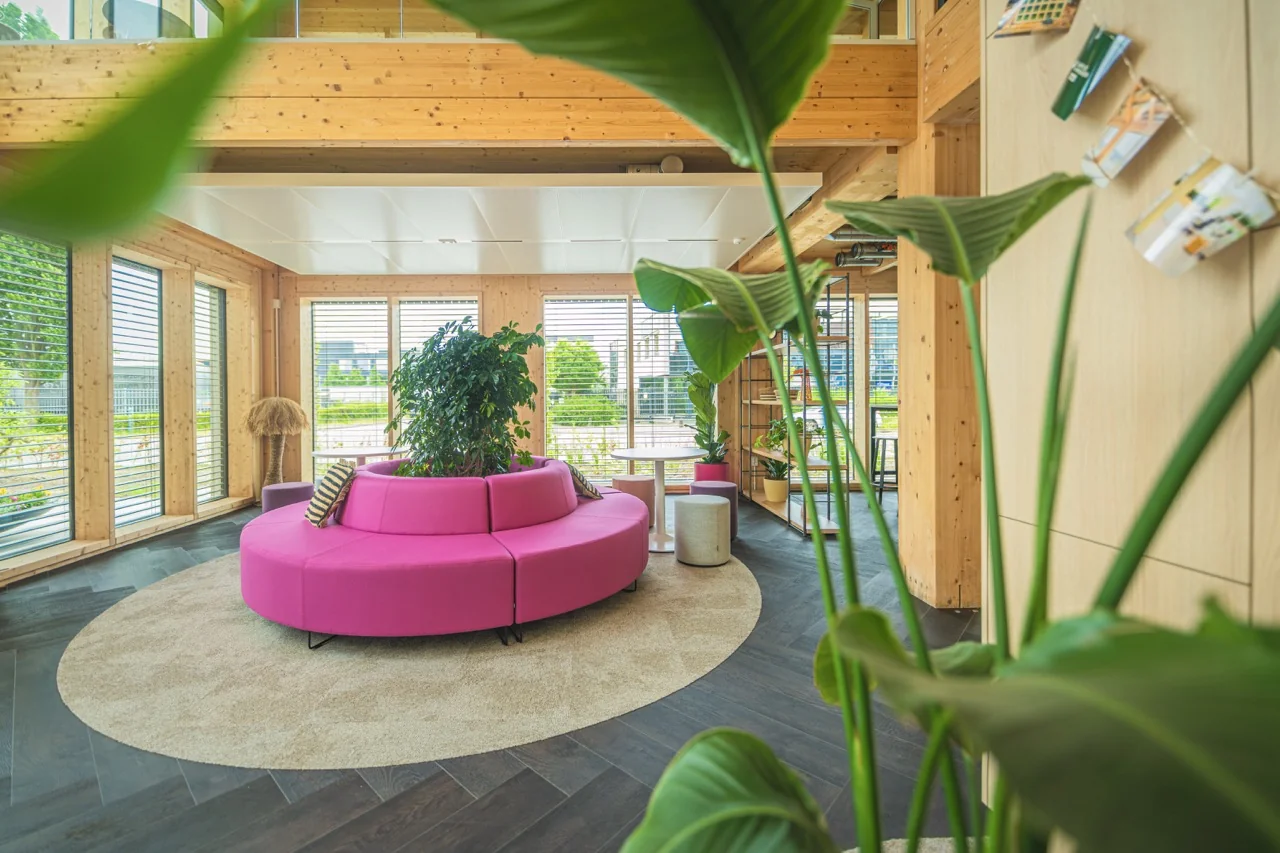 Moderne loungehoek met een ronde roze bank, omringd door planten en grote ramen in een houten interieur.