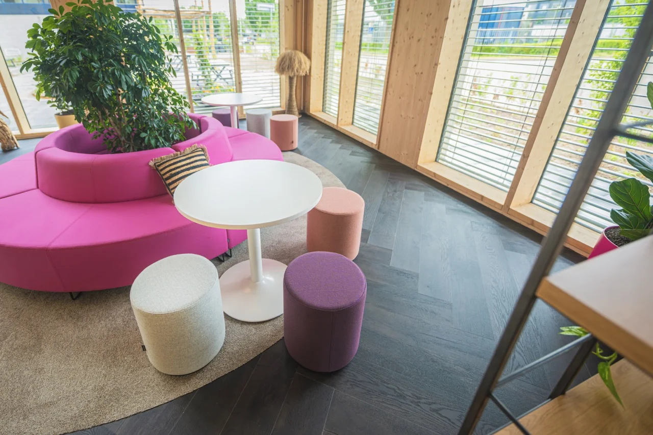 Moderne loungehoek met roze ronde zitbank, kleurrijke krukjes en een witte tafel in een houten interieur met veel daglicht.