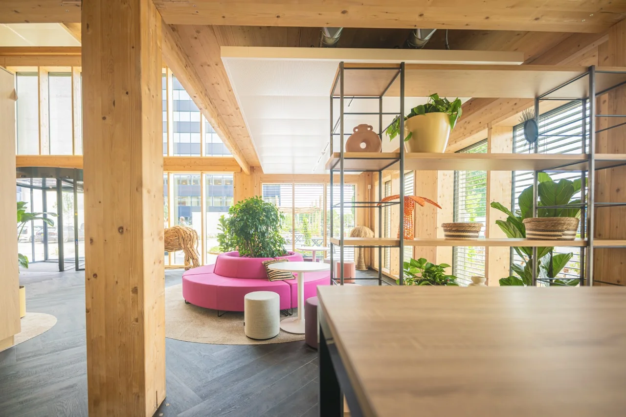 Lichte en moderne zithoek met een ronde roze bank, veel groen en houten interieurdetails in een open kantoorruimte.