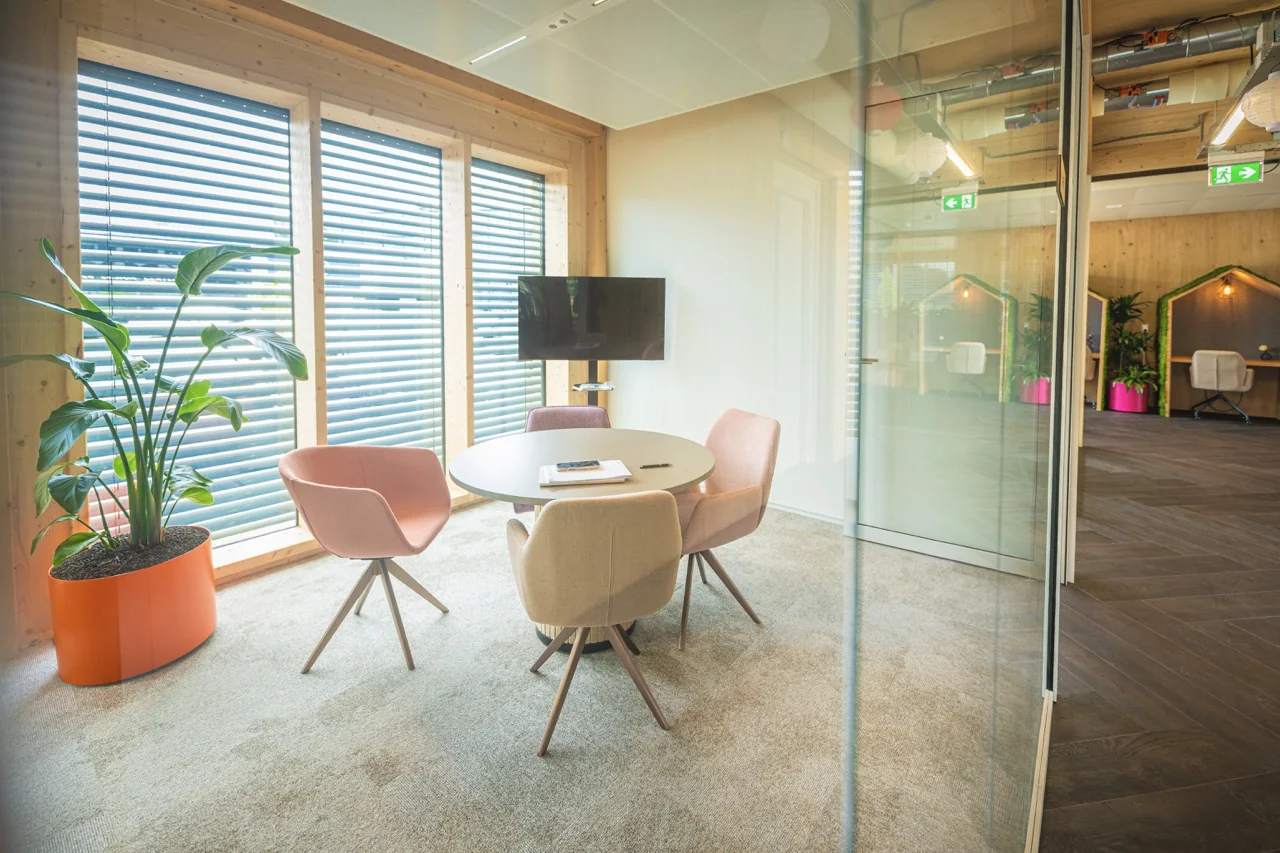Moderne vergaderruimte met ronde tafel, pastelgekleurde stoelen, flatscreen en grote ramen met jaloezieën.