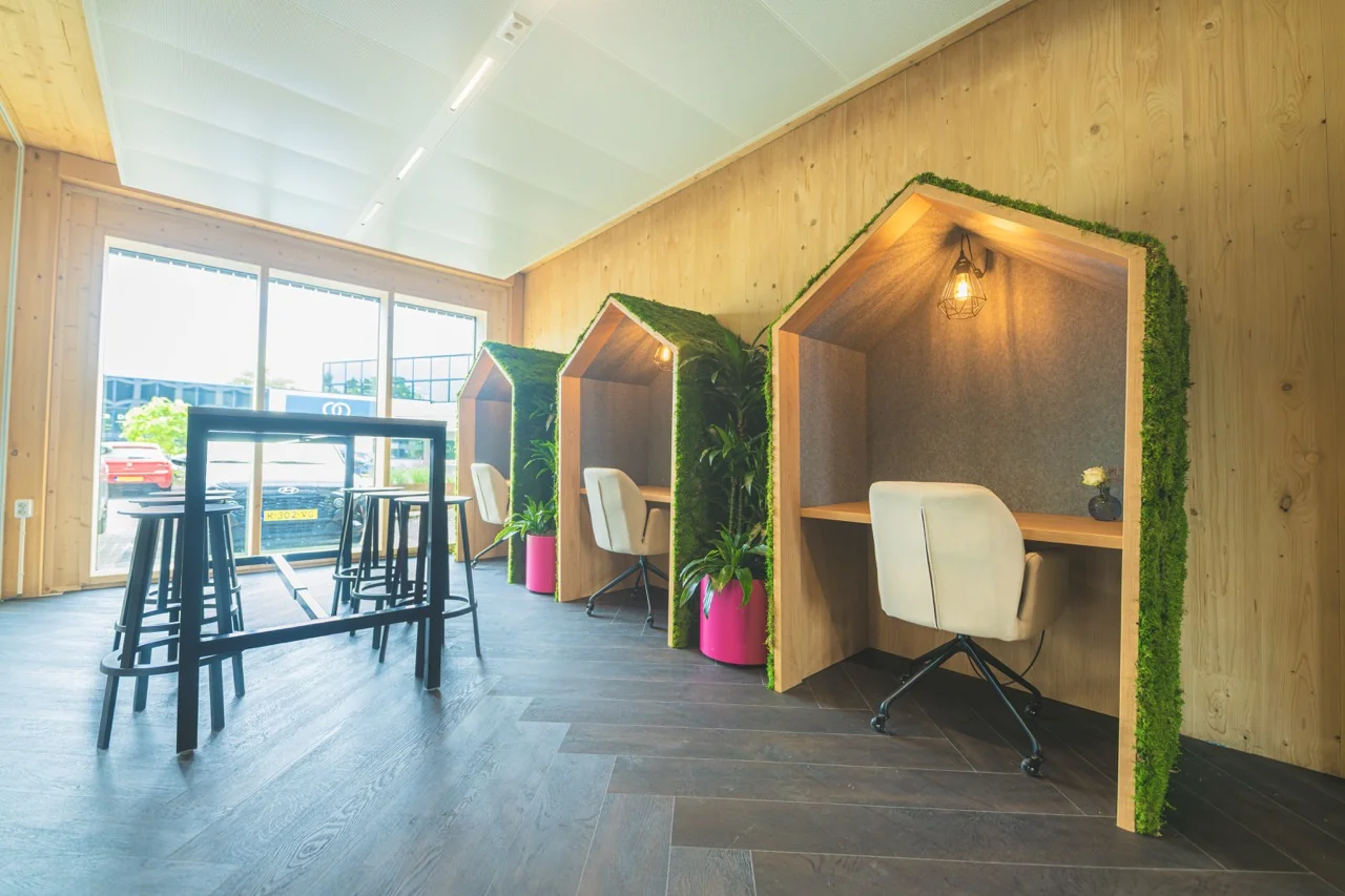 Moderne werkplek met houten belhokjes in huisvorm, omgeven door planten en voorzien van bureaustoelen.