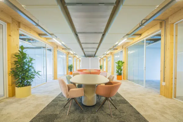 Moderne kantoorruimte met houten accenten, glazen wanden, kleurrijke stoelen en planten langs een centrale vergadertafel.