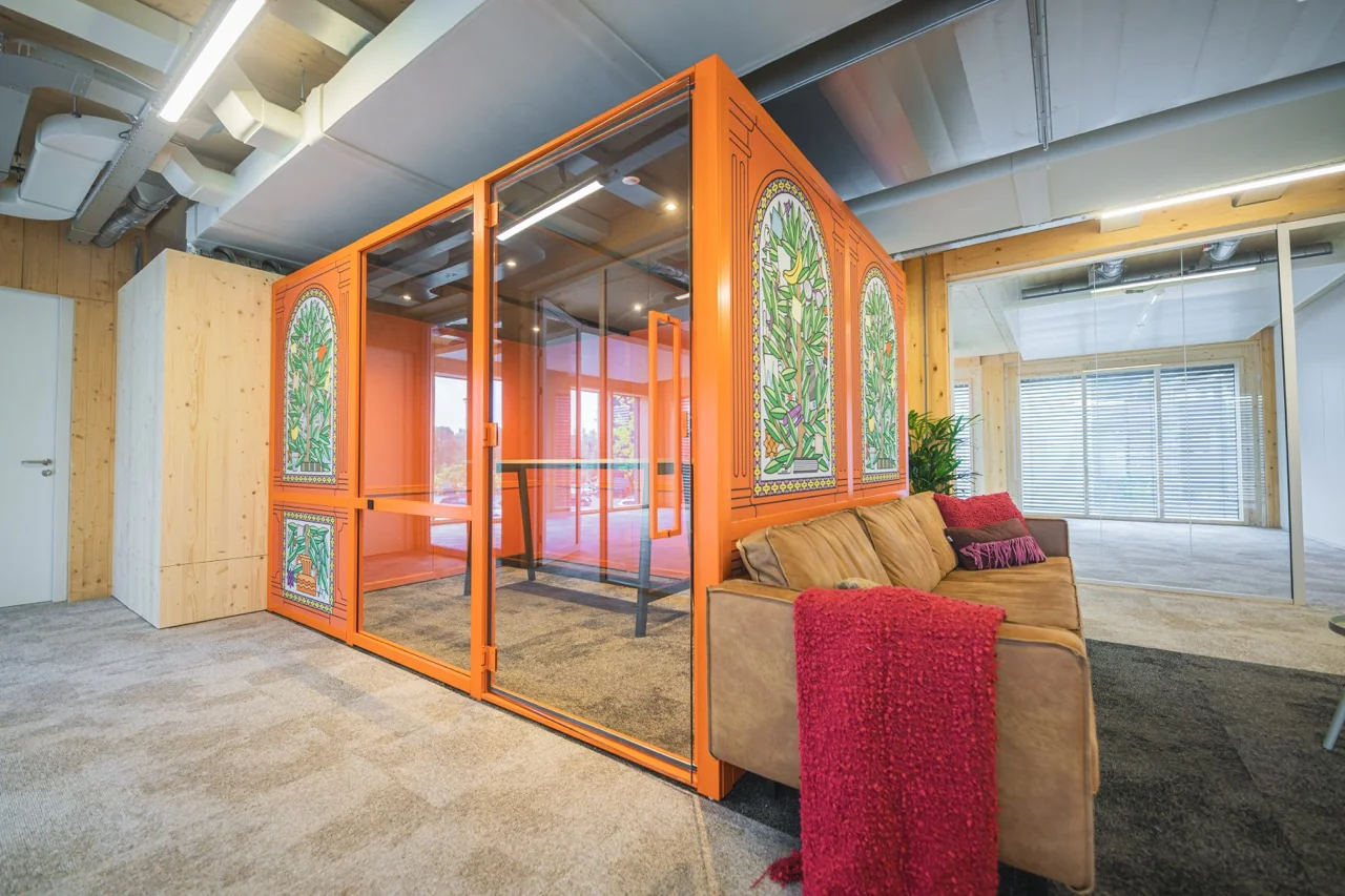 Moderne kantoorruimte met een feloranje vergaderpod voorzien van kleurrijke glasschilderingen en een comfortabele zithoek met een bruine bank en rode plaid.