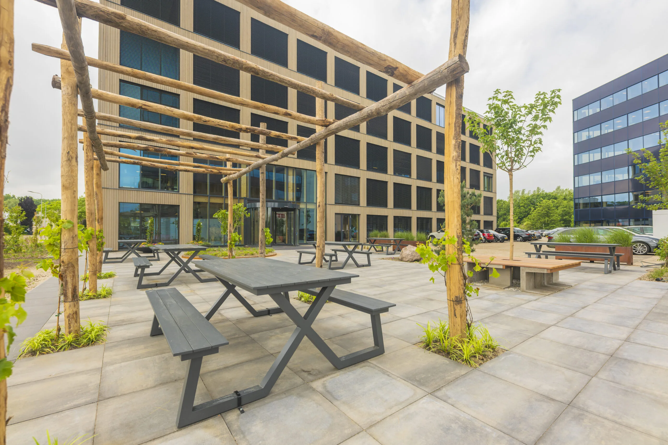 Moderne buitenruimte met houten pergola, picknicktafels en jonge beplanting bij een kantoorgebouw.