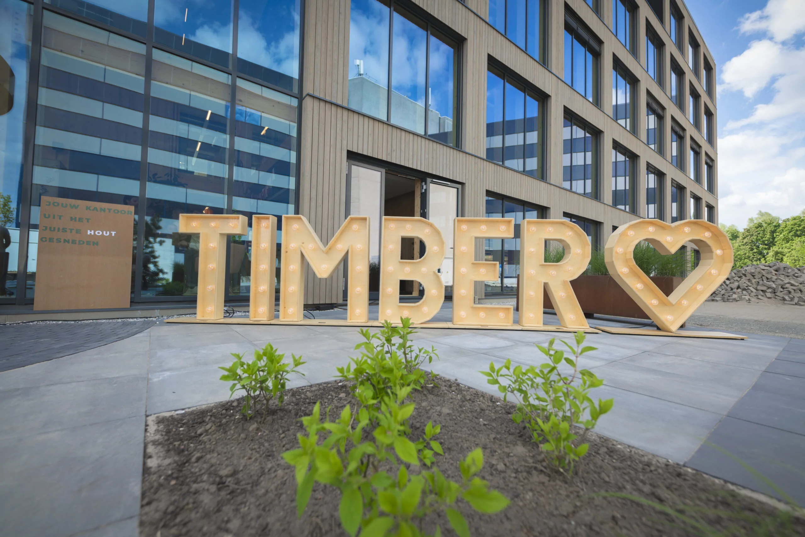 Grote houten lichtletters vormen het woord "TIMBER" met een hartsymbool, geplaatst voor een modern houten kantoorgebouw.
