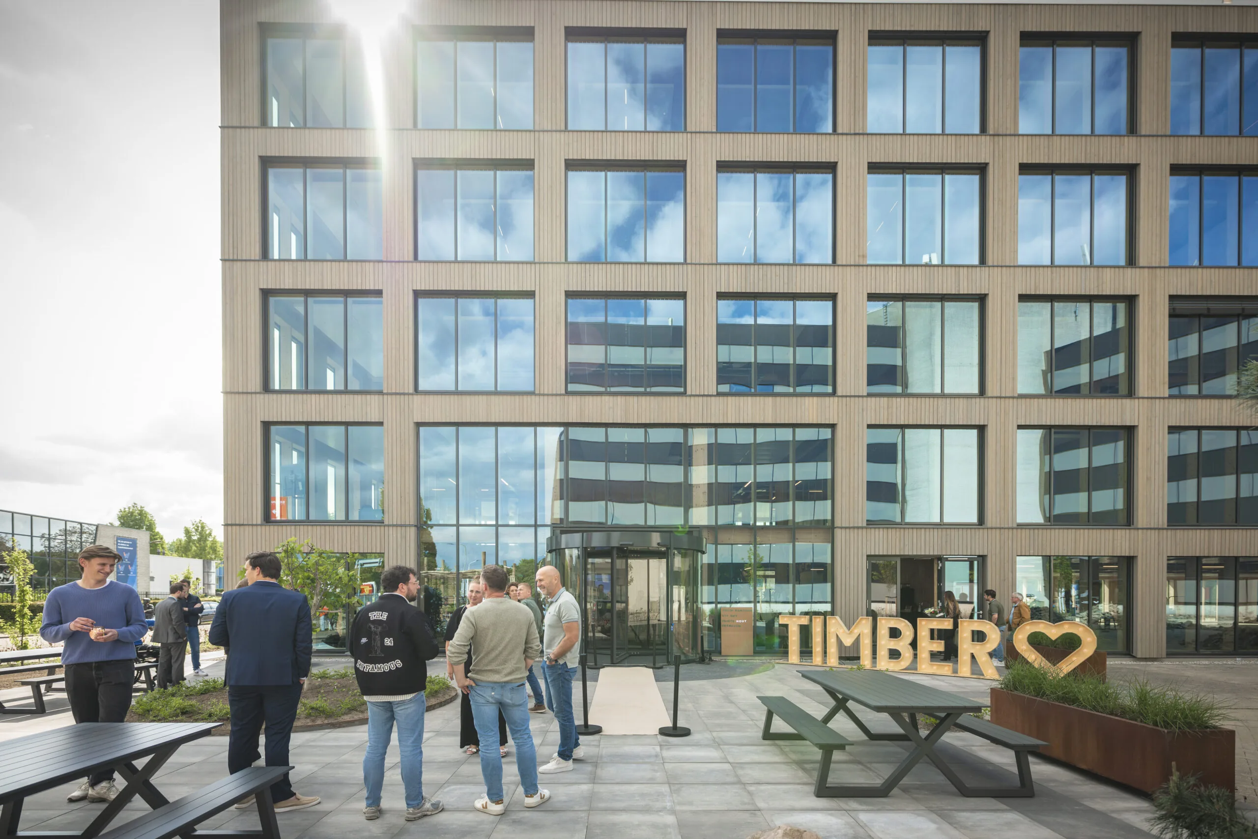 Groep mensen verzameld op een modern plein voor het houten kantoorgebouw TIMBER met grote ramen en zonlicht op de achtergrond.