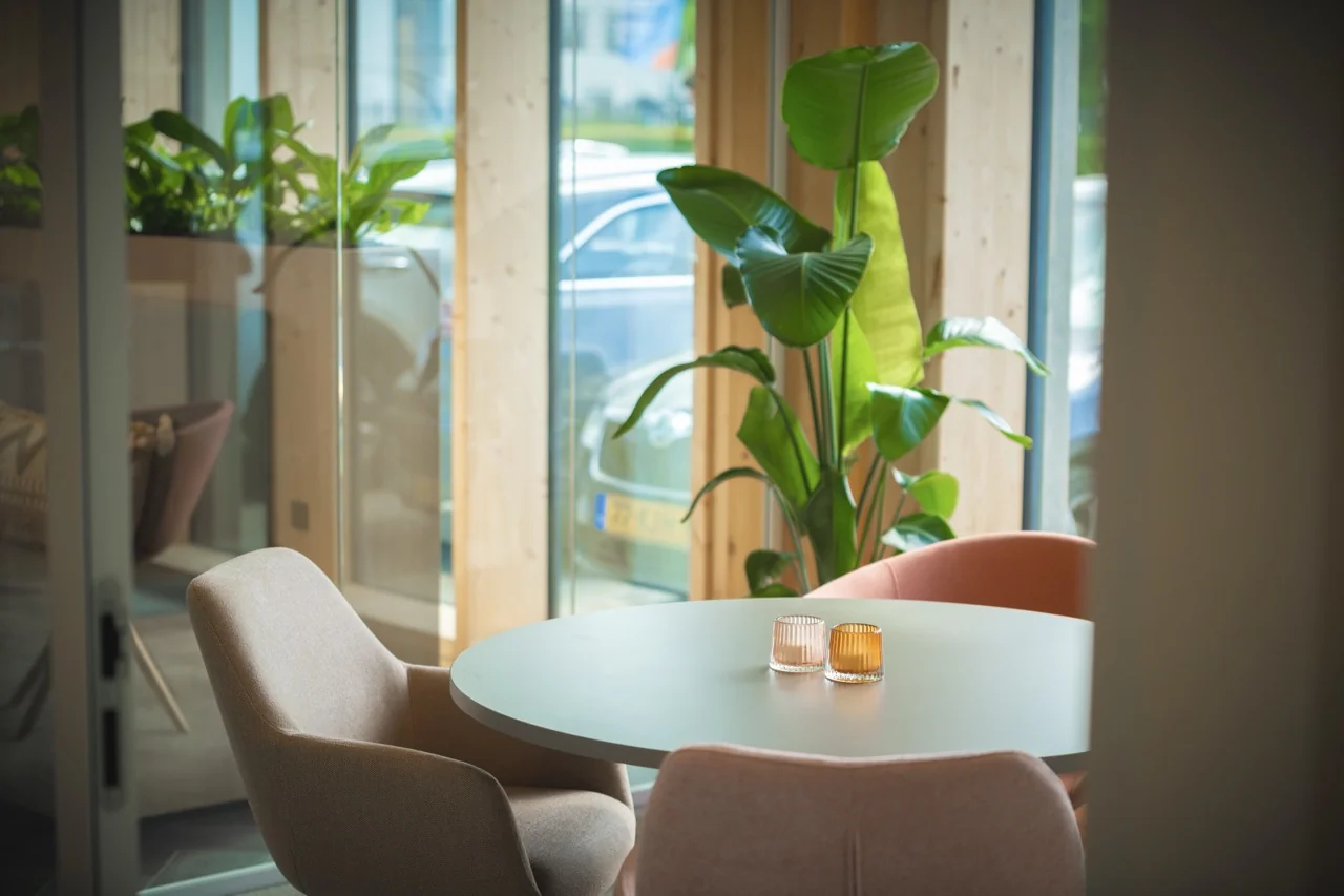 Ronde tafel met vier moderne stoelen, twee gekleurde kaarsen en een grote kamerplant bij een glazen wand.