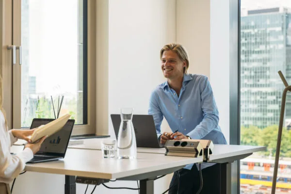 Man in blauwe blouse voert een ontspannen gesprek aan een bureau in een modern kantoor met grote ramen.