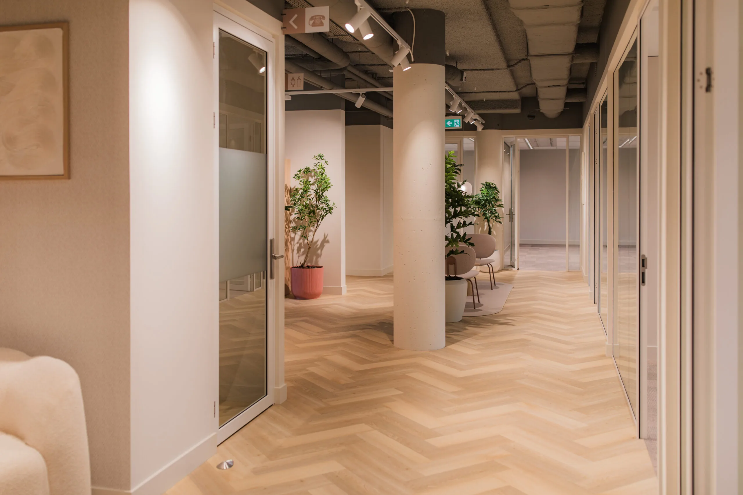 Modern office hallway in Hullenberg 143 with wooden herringbone flooring, glass doors, potted plants, and neutral-toned decor.
