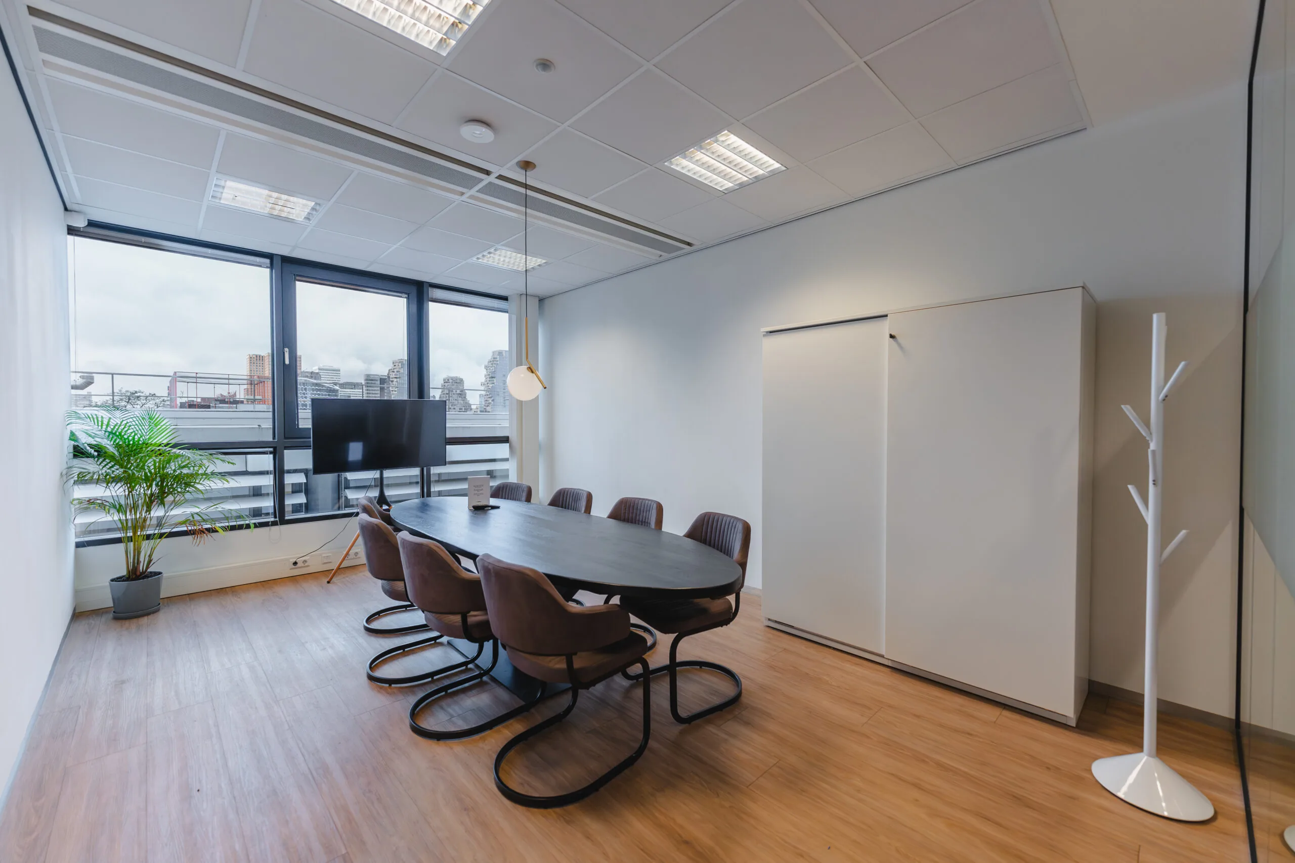 Moderne vergaderruimte met ovale tafel, acht stoelen, een flatscreen en uitzicht op een stedelijk landschap.