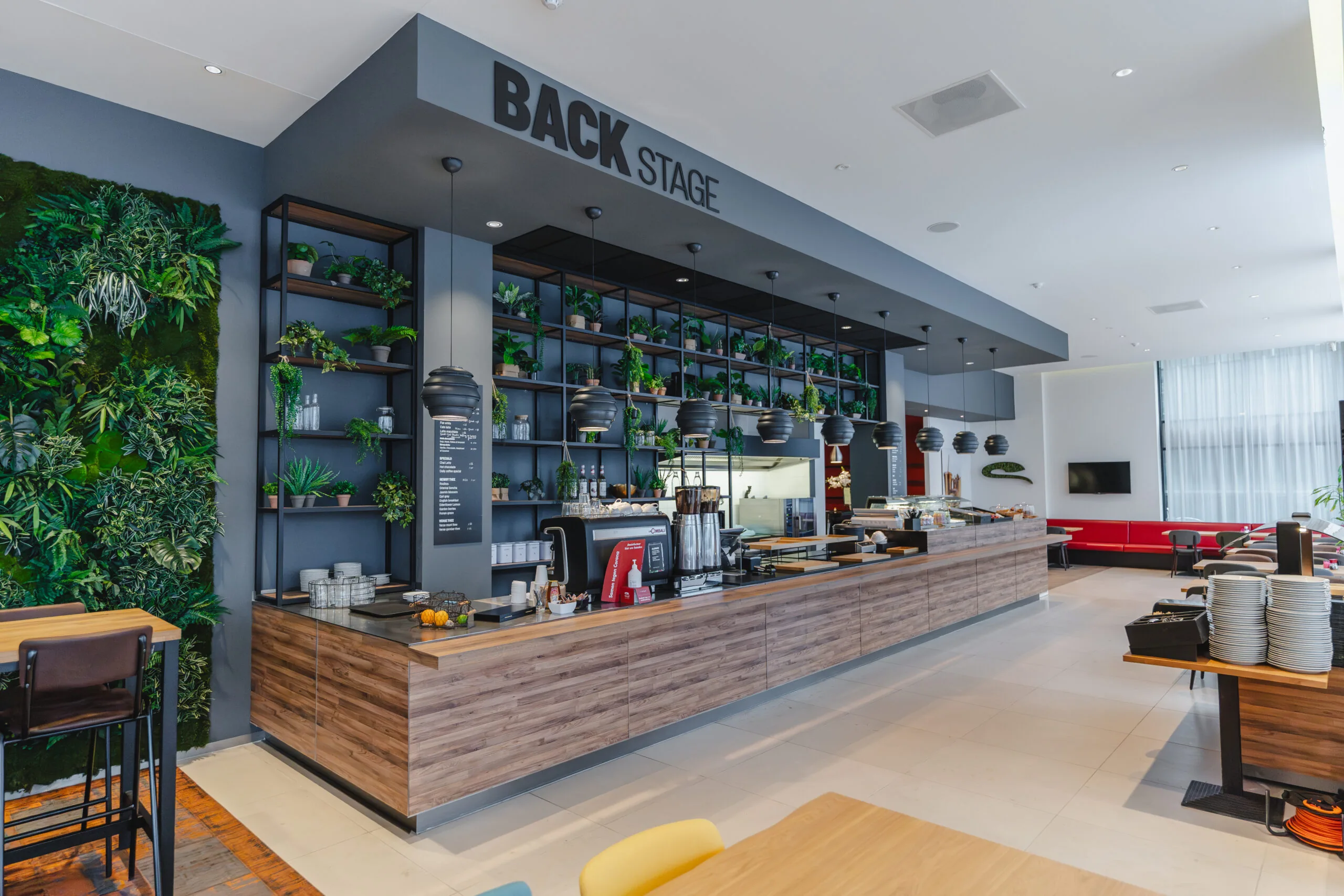 Moderne kantine met houten bar, groene plantendecoratie en de tekst "BACKSTAGE" boven de uitgiftebalie.