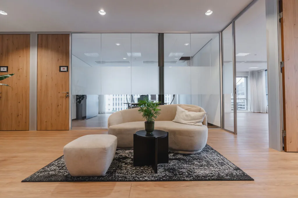 Moderne gangruimte met een beige zitbank, een poef, een zwart bijzettafeltje en een plant op een vloerkleed, omgeven door houten deuren en glazen wanden.