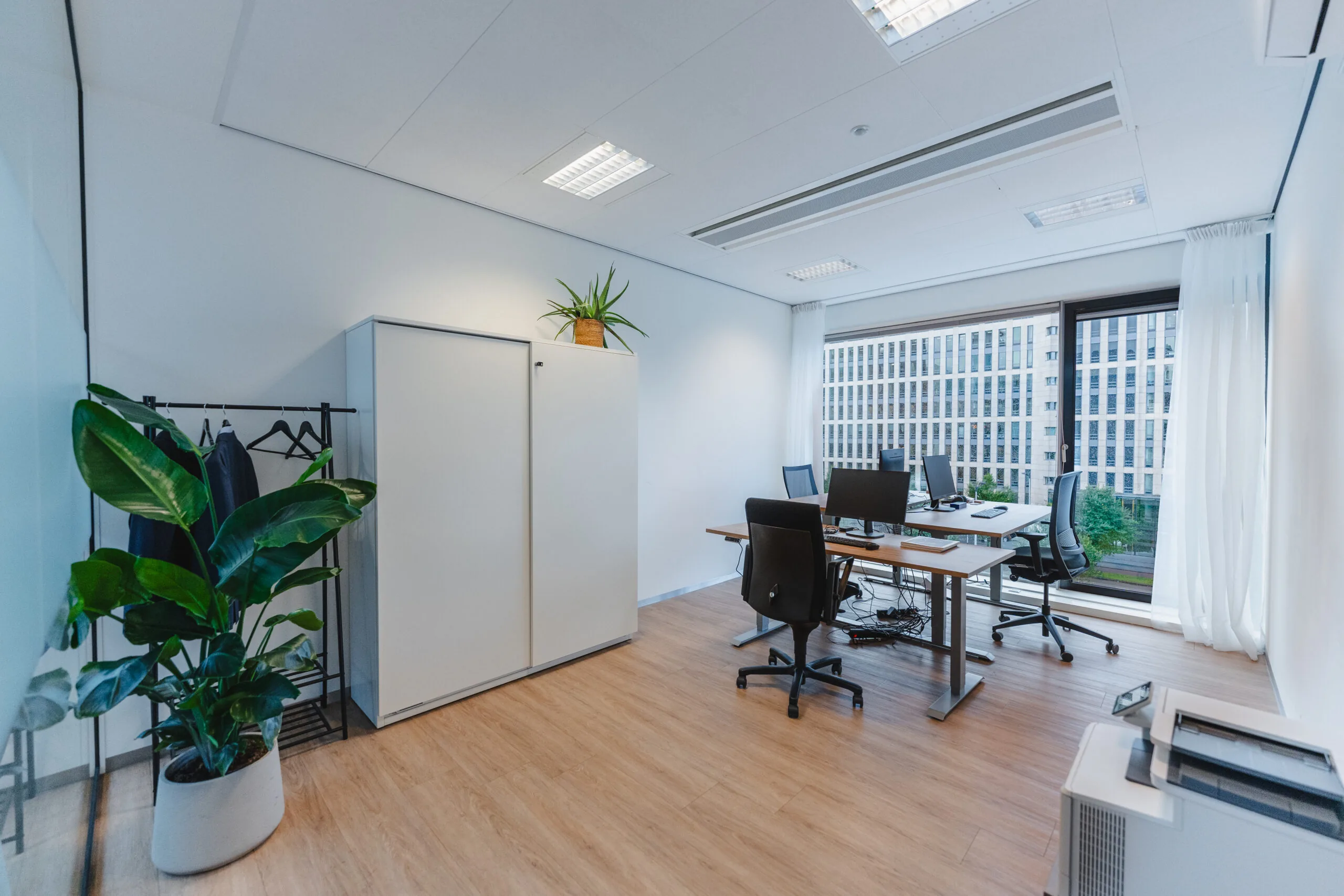 Moderne kantoorruimte met bureaus, computers, planten en uitzicht op een kantoorgebouw.