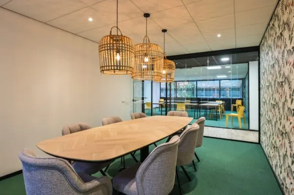 Vergaderruimte aan de Havenweg 24 met een ovale houten tafel, gestoffeerde stoelen en rotan hanglampen, grenzend aan een ruimte met gele stoelen achter een glazen wand.