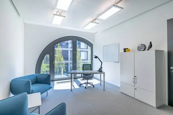 Modern kantoor met groot boograam, een bureau, blauwe fauteuils en een witte kast.