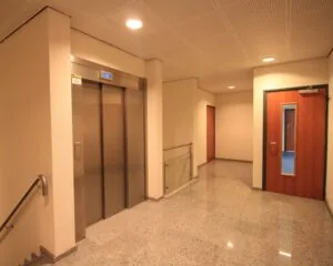 Verlichte binnenhal met een gesloten liftdeur, twee houten deuren en een trapleuning.