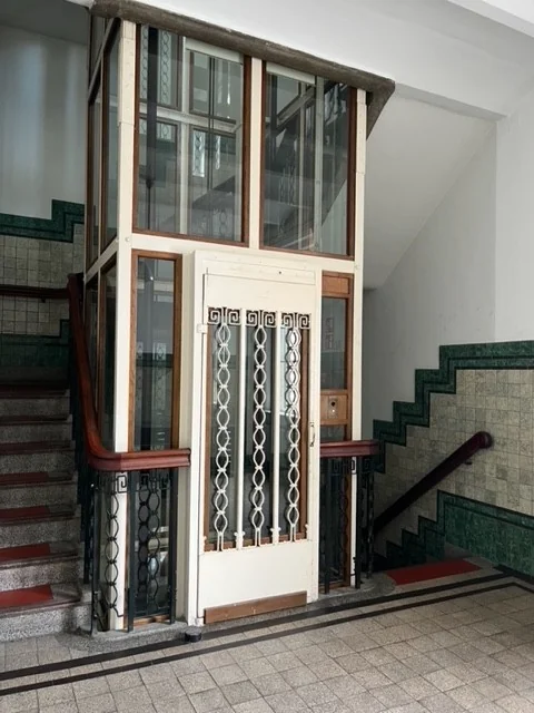 Een vintage lift met glazen panelen en decoratief hekwerk bevindt zich in een trapportaal.