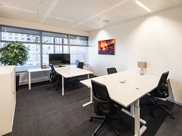 Een moderne kantoorruimte met vier bureaus, een plant, en een schilderij aan de muur.