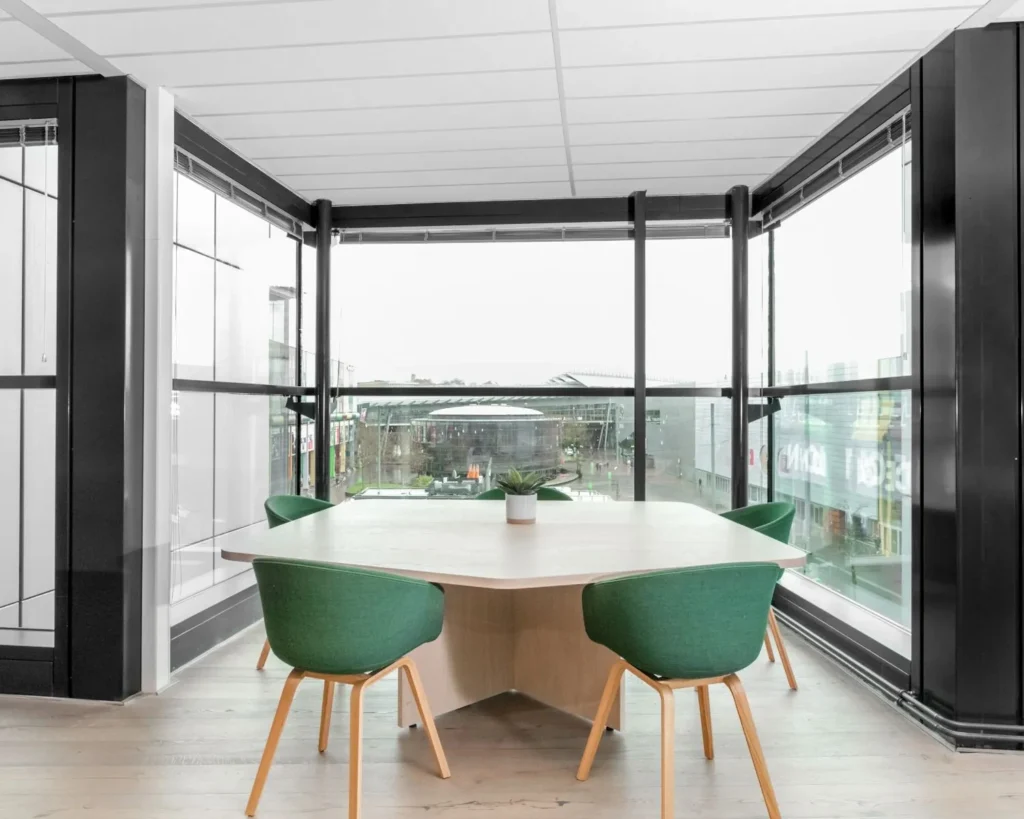 Een moderne kantoorruimte met een grote tafel en groene stoelen bij een raam met uitzicht op de Johan Cruijff Boulevard.