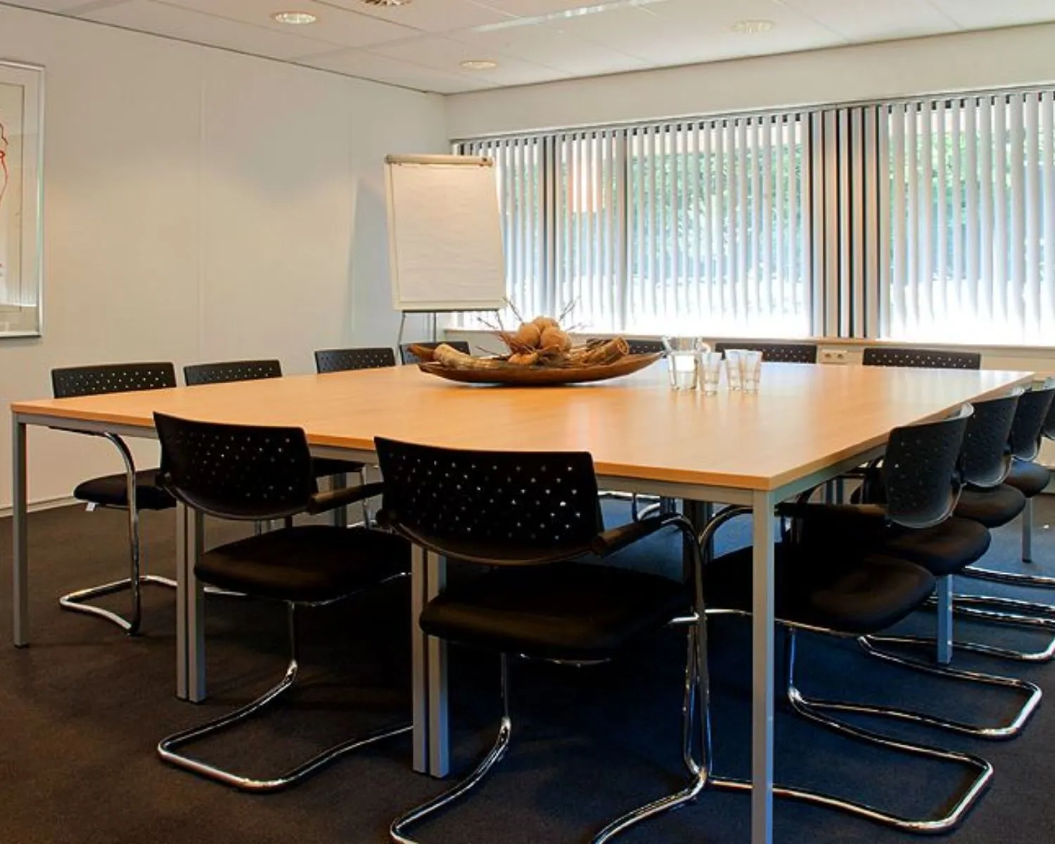Vergaderruimte met een vierkante tafel, twaalf stoelen, flip-over en grote ramen met verticale lamellen in een kantoor aan de Herenweg 15 in Heemstede.