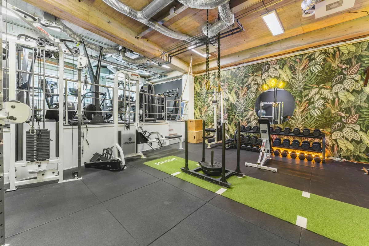 Een moderne fitnessruimte met diverse trainingsapparaten en een plantenmotief op de muur.