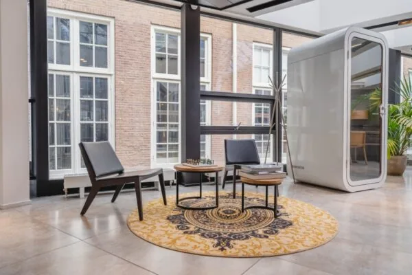 Een moderne kantoorruimte in Amsterdam met strakke stoelen, een ronde decoratieve tapijt en een glazen cabine naast grote ramen.