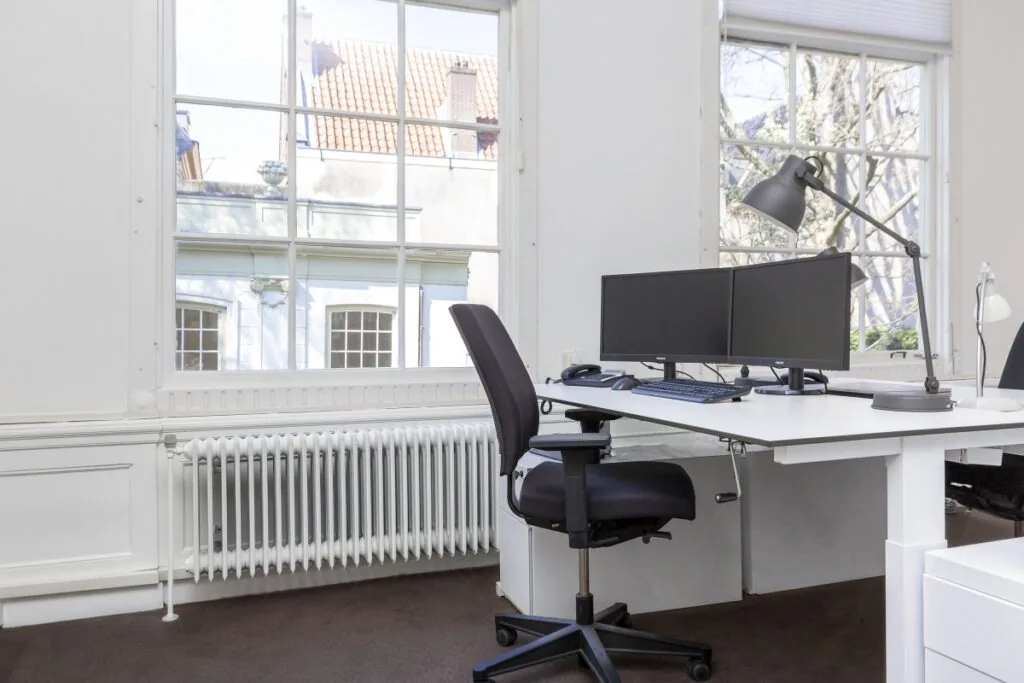 Modern kantoor met dubbele monitoren en bureaustoel bij een groot raam aan de Keizersgracht 64.