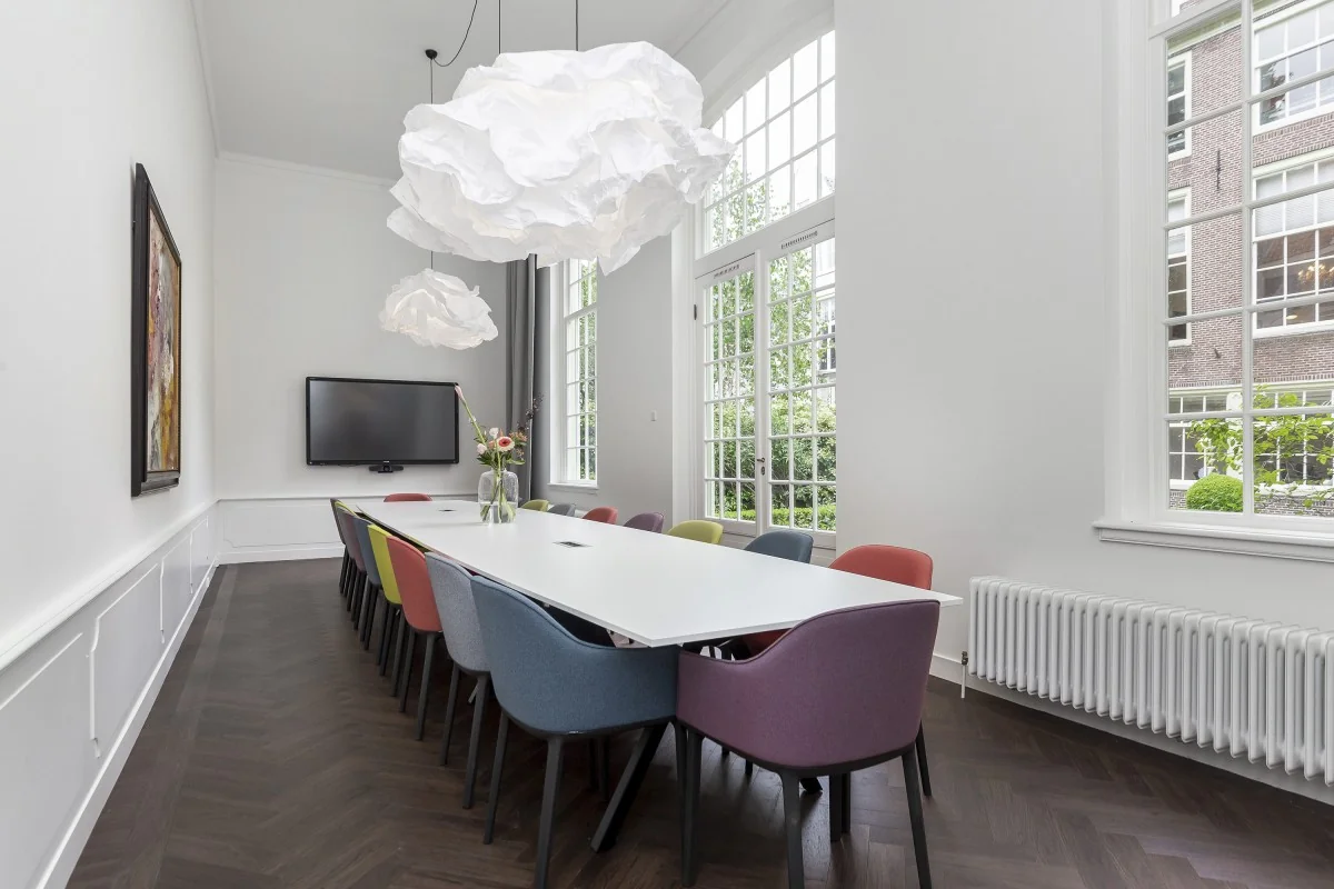 Vergaderruimte in Keizersgracht 64 met lange witte tafel, kleurrijke stoelen, grote ramen en speelse hanglampen.