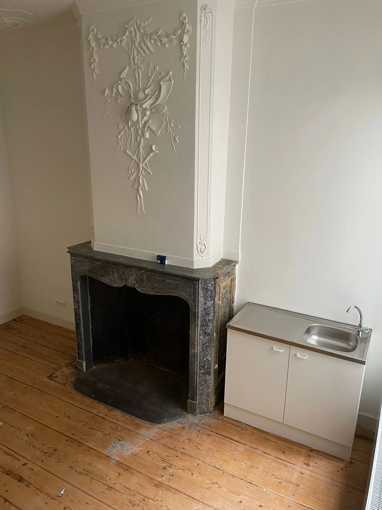 Een klassieke schouw met sierlijk stucwerk naast een eenvoudig wit keukenkastje met spoelbak in een kamer aan de Kloveniersburgwal 56.