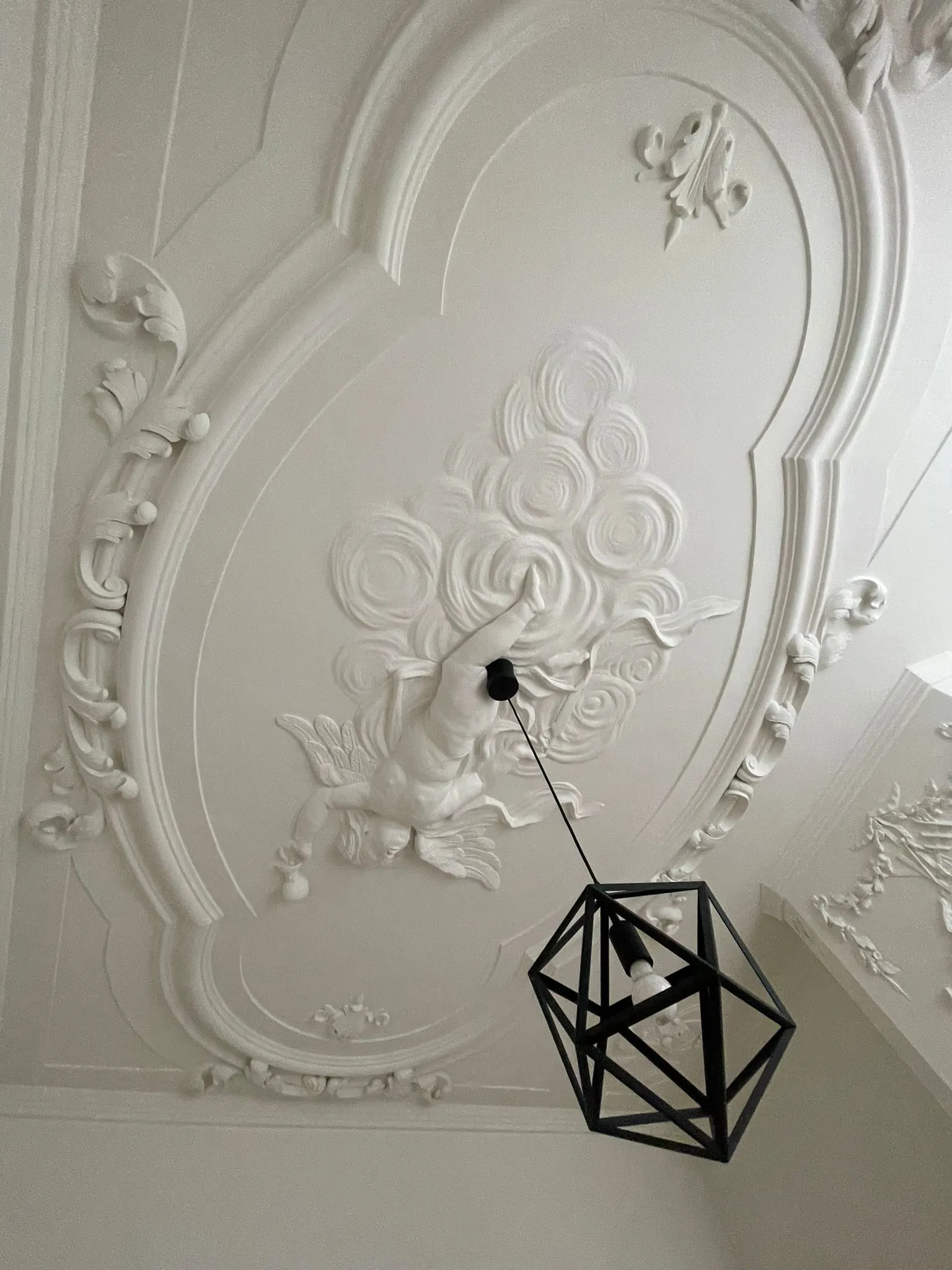 Plafondornament met sierlijke stucwerkdecoratie en engelfiguur aan het plafond van Kloveniersburgwal 56, met een moderne zwarte hanglamp.