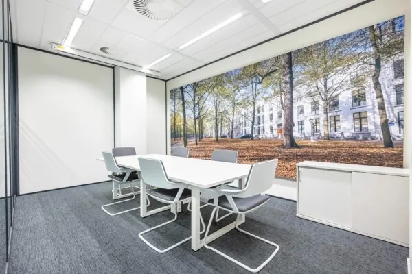 Moderne vergaderruimte met witte tafel, grijze stoelen en een grote wandfoto van het Koningin Julianaplein in de herfst.