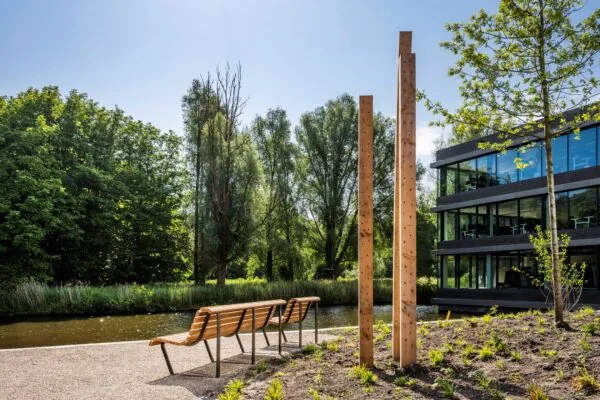 Bankjes aan het water bij een modern kantoorgebouw aan de Nachtwachtlaan, omgeven door bomen en kunstzinnige houten palen.